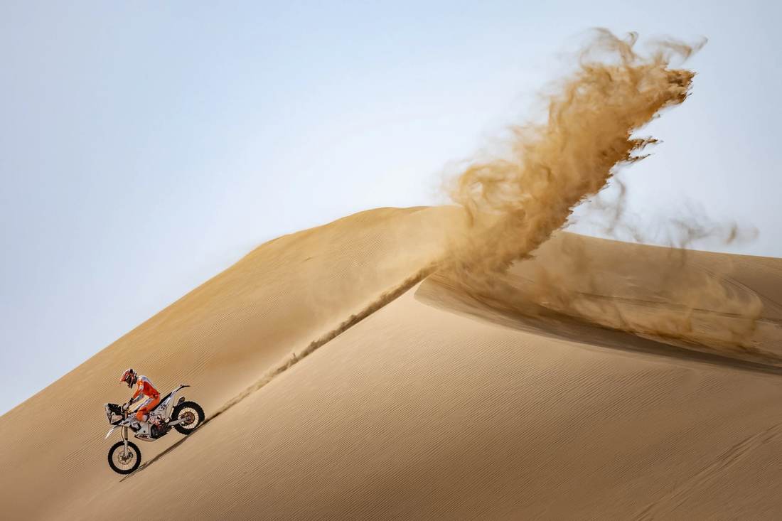 Rally motorcycle cresting a dune, throwing a plume of sand