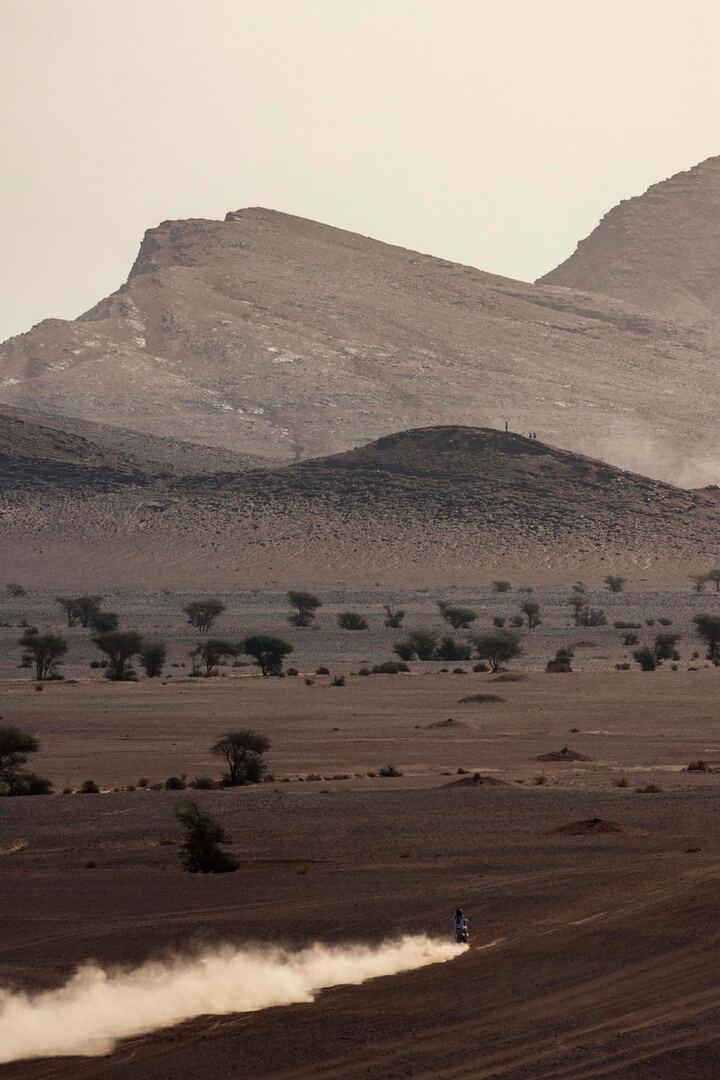 A distant motorcycle leaves a long dust trail across a desert valley