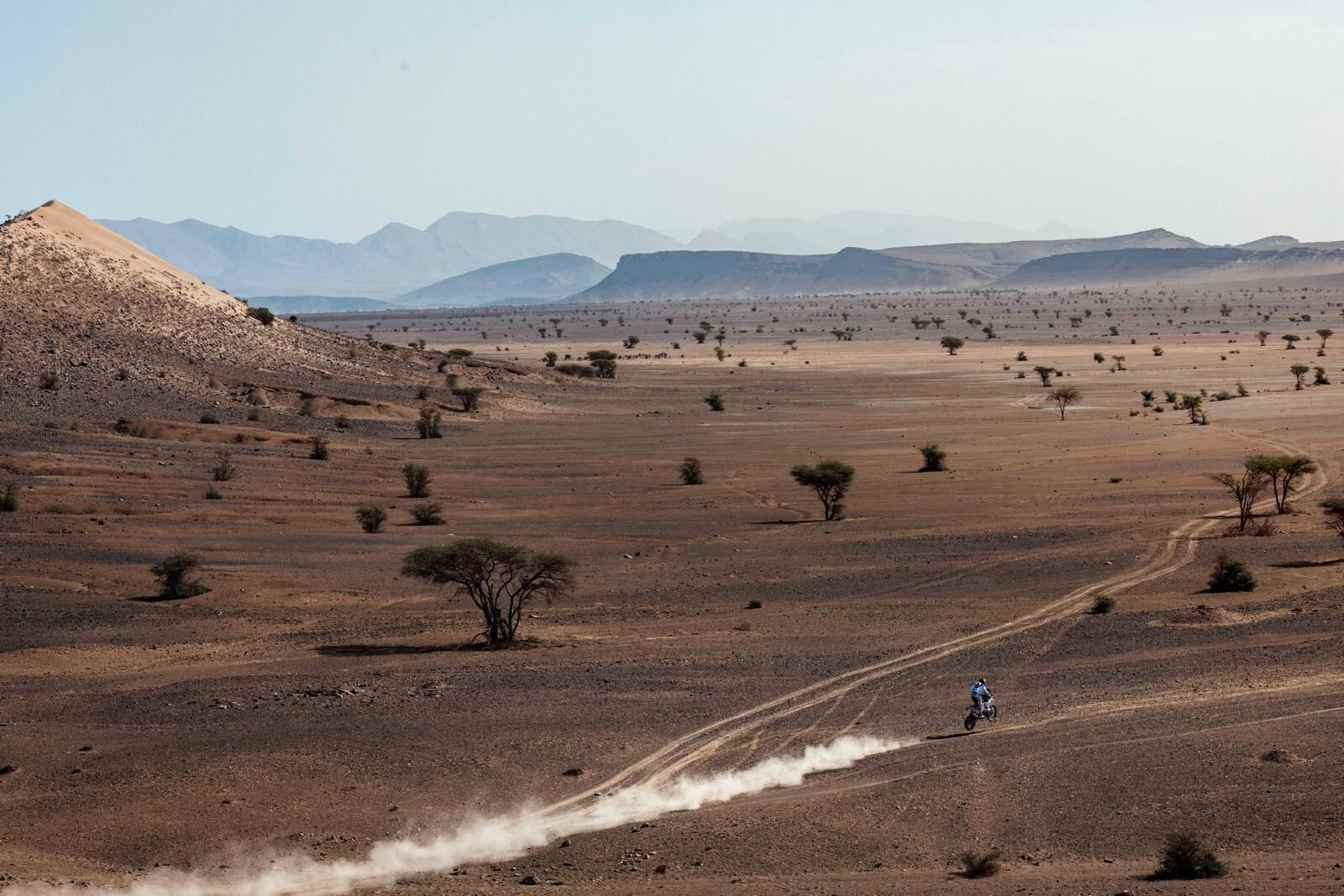 A motorcycle crosses open desert ground, trailing dust below rocky hills