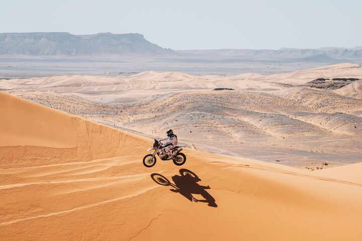 A rally motorcycle crosses a dune crest above a desert valley